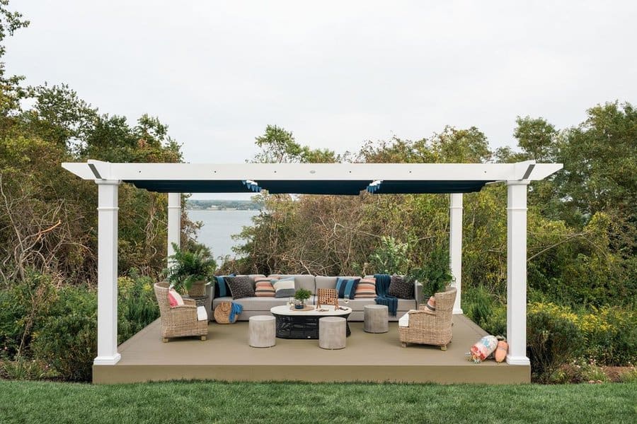 Scenic outdoor lounge with a white pergola, cozy seating, colorful cushions, and a fire pit overlooking lush greenery and a lake