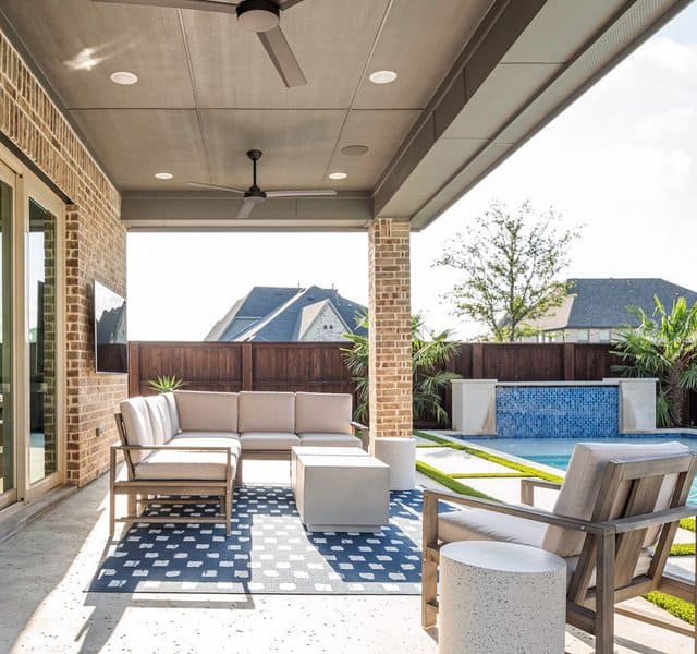 Modern covered patio with neutral furniture, ceiling fans, a geometric rug, and a poolside view framed by a wooden fence