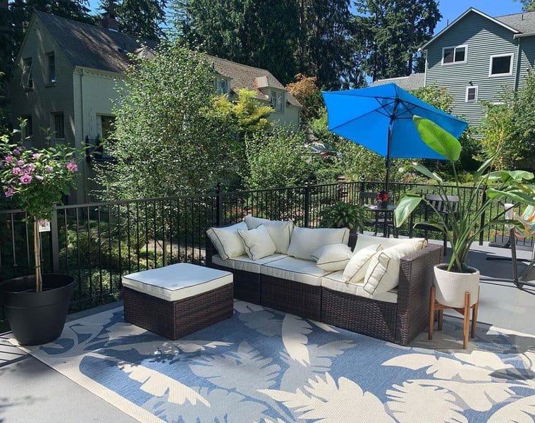 Bright outdoor patio with wicker furniture, cream cushions, a blue umbrella, potted plants, and a leafy patterned rug