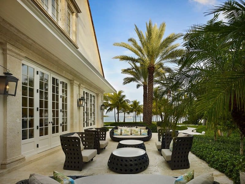 Elegant outdoor patio with modern black furniture, light cushions, and palm trees overlooking a serene waterfront view, creating a luxurious tropical retreat