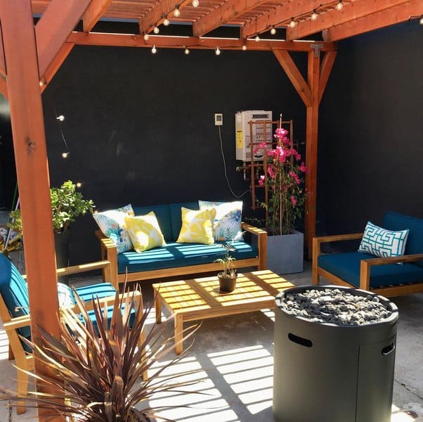 Cozy pergola-covered patio with wooden furniture, teal cushions, vibrant throw pillows, string lights, and blooming pink flowers