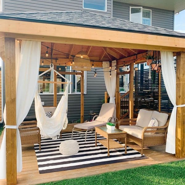 Charming gazebo with wicker furniture, a hammock chair, striped rug, string lights, and flowing white curtains for a cozy retreat