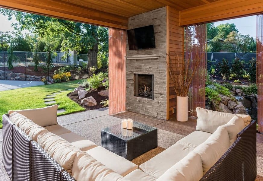 Modern covered patio with a stone fireplace, cozy sectional seating, and a serene garden view for year-round comfort