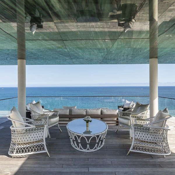 Elegant outdoor seating area with white wicker furniture, glass table, and breathtaking ocean views under a modern shaded canopy