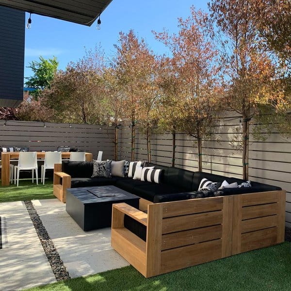 Outdoor patio with wooden furniture, a black sofa, dining table, and trees lining the fence