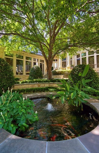 Circular koi pond with a small fountain, surrounded by lush plants and shaded by a large tree in a garden.