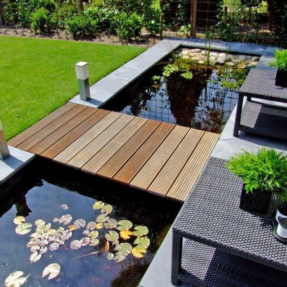 Small wooden bridge over a pond with water lilies, surrounded by patio seating and greenery.