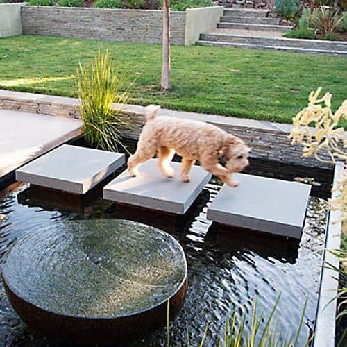 Floating concrete steps over a pond with a circular water feature, surrounded by greenery and landscaping.