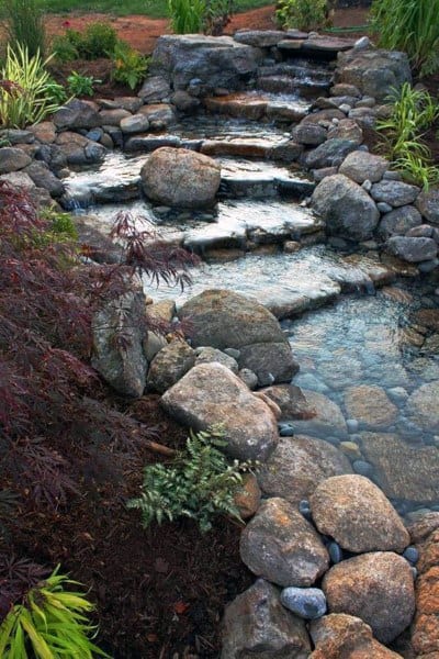 Natural tiered pond with cascading water over rocks, surrounded by greenery and ornamental plants.