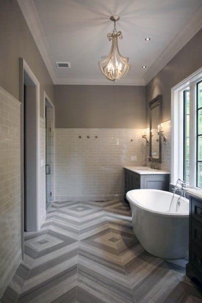 Spacious bathroom with freestanding tub, large window, chevron tile floor, and chandelier.