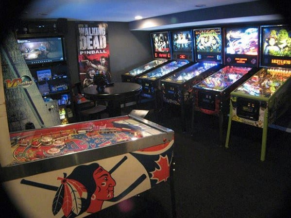 A room filled with various pinball machines, including a Walking Dead-themed one, in a dimly lit arcade