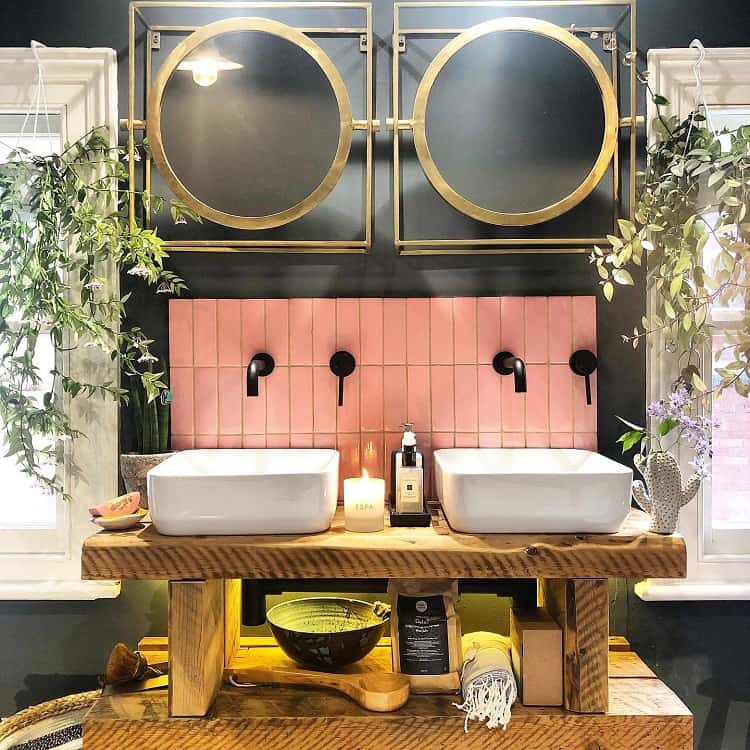 Double vanity with pink tile backsplash, gold-framed mirrors, and wooden shelf in a stylish bathroom.