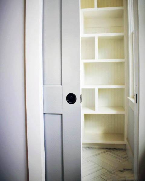 White pocket door with a black handle leads to a pantry with built-in cream shelves and herringbone-patterned flooring