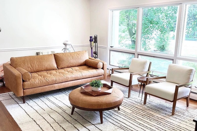 Bright living room with tan leather sofa, white armchairs, round ottoman, and large windows.