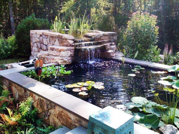 Stone waterfall feature flowing into a pond with water lilies, surrounded by greenery in a garden.