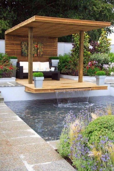 Elevated wooden deck with a pergola, overlooking a pebble-bottom pond with dual waterfall features.