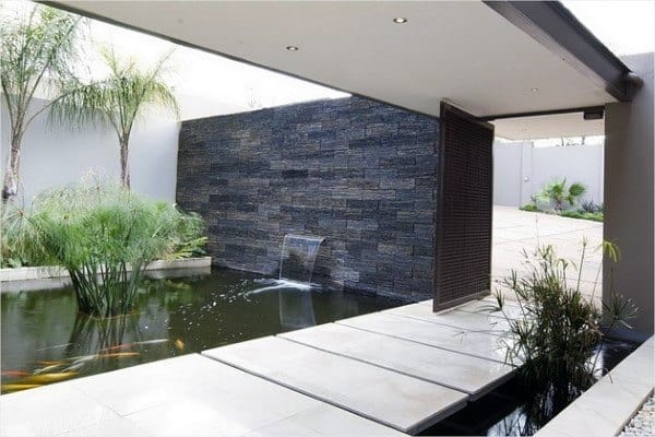 Modern deck pond with floating stone steps, a sleek waterfall, and aquatic plants, surrounded by clean landscaping.