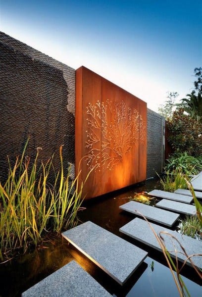 Floating stone steps over a pond with artistic lighting, featuring a decorative metal wall and tall plants.