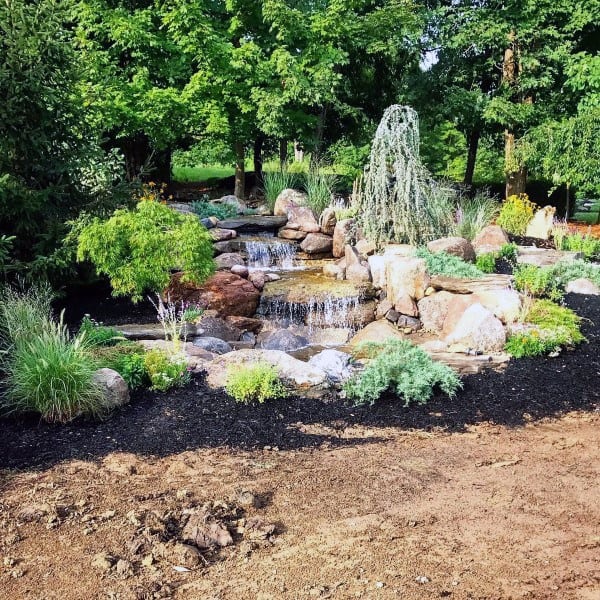 Natural tiered pond with cascading waterfalls, surrounded by lush greenery, rocks, and ornamental plants.