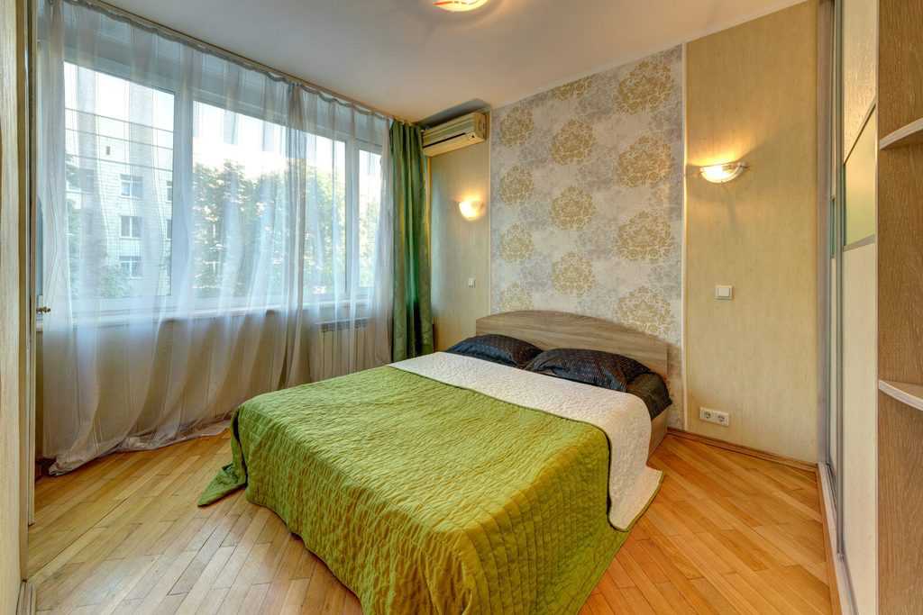 Bedroom with floral wallpaper, green bedding, sheer curtains, and wood flooring.