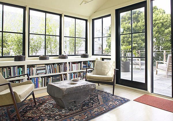 A cozy sunroom with a built-in bookshelf, minimalist armchairs, a rustic stone coffee table, and black-framed windows.