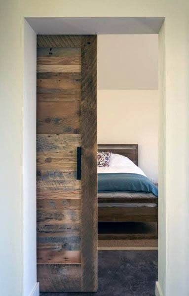 Rustic reclaimed barn wood pocket door with a black handle opens to a cozy bedroom featuring a wooden bed and soft bedding