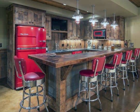 Rustic garage bar with red retro fridge, wooden cabinets, red stools, and industrial lighting.