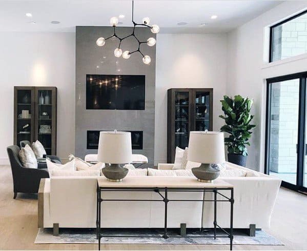 Modern living room with white sofas, a large TV, a geometric chandelier, and floor-to-ceiling windows