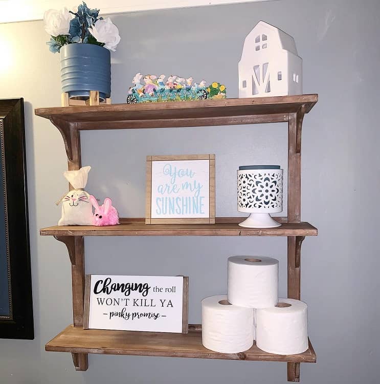 Rustic wooden bathroom shelf with decor, toilet rolls, and motivational signs.