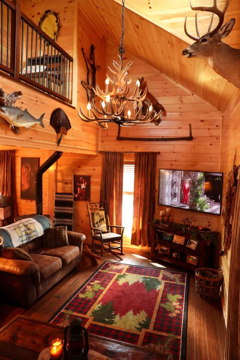 Cozy cabin-style living room with brown wood paneling, antler chandelier, rustic decor, and mounted deer head