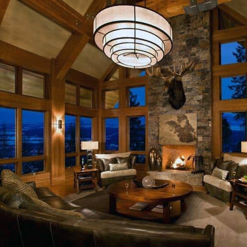 A rustic chic living room featuring a stone fireplace, a striking chandelier, leather seating, and large windows with a scenic view, creating a cozy and sophisticated atmosphere