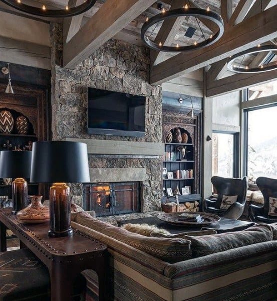 A rustic contemporary living room featuring a stone fireplace, exposed beams, modern lighting, and large windows that offer a stunning view, creating a cozy yet stylish atmosphere