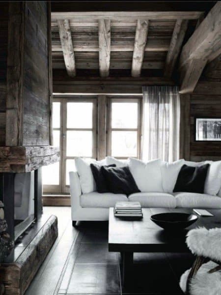 A rustic country living room featuring a sleek white sofa, wooden beams, a stone fireplace, and large windows, creating a cozy yet modern atmosphere