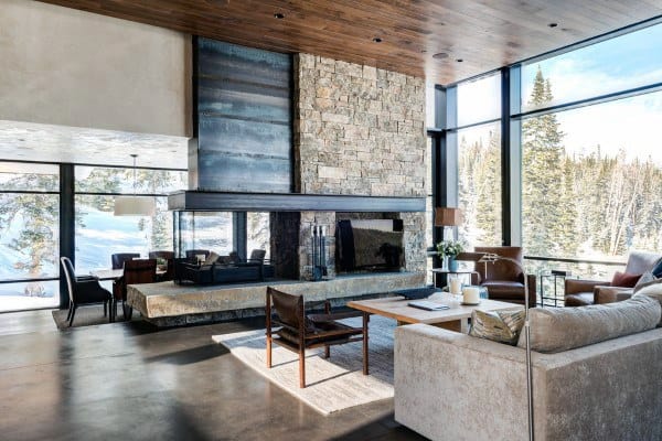 A modern rustic living room with a stone fireplace, large windows offering panoramic mountain views, and contemporary furnishings, creating a sleek yet cozy atmosphere