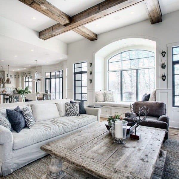 A rustic farmhouse living room with exposed wooden beams, a cozy sofa, a vintage wood coffee table, and large windows that let in natural light, creating a bright and inviting space