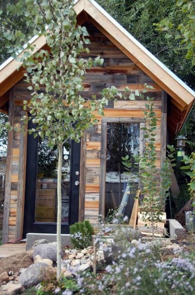 Rustic wood shed with glass door and large windows, surrounded by trees and plants.