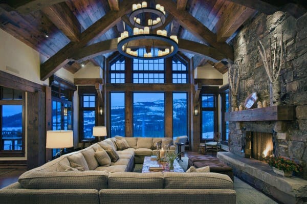 A spacious rustic living room with a stone fireplace, exposed wooden beams, large windows showcasing scenic mountain views, and cozy seating, creating a warm and inviting atmosphere