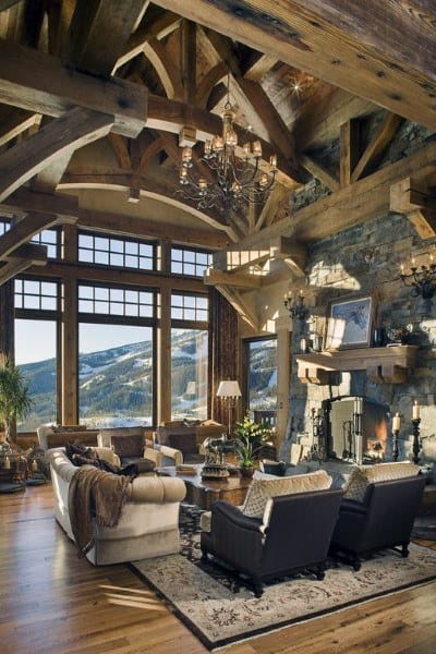 A rustic living room featuring exposed wooden beams, a stone fireplace, large windows with stunning mountain views, and elegant furniture, creating a luxurious yet cozy atmosphere