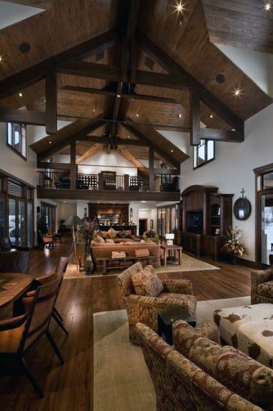 A spacious rustic living room with vaulted wooden beams, large windows, comfortable seating, and an open loft area, creating an airy and welcoming atmosphere