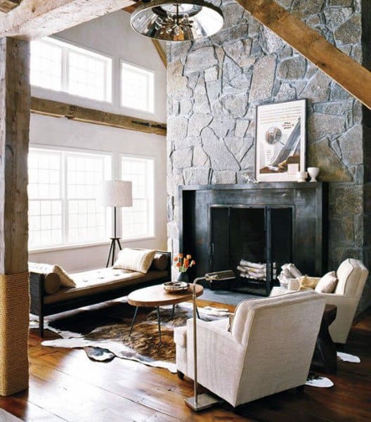 A rustic living room with a stone fireplace, wooden beams, modern furniture, and large windows, creating a cozy, inviting atmosphere