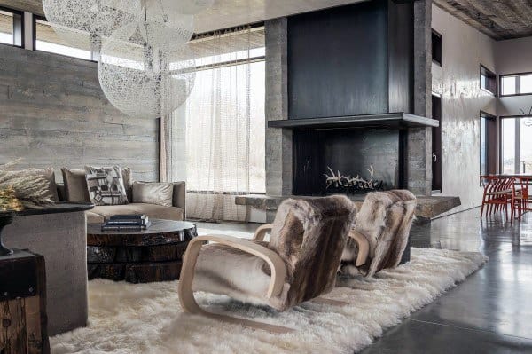 A modern rustic living room featuring a sleek fireplace, fur chairs, a cozy rug, and large windows offering scenic views, creating a warm, minimalist space