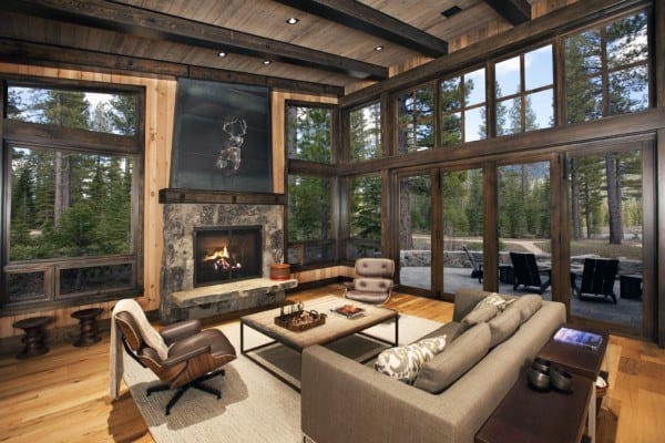 Cozy rustic living room with large windows, a stone fireplace, and forest views