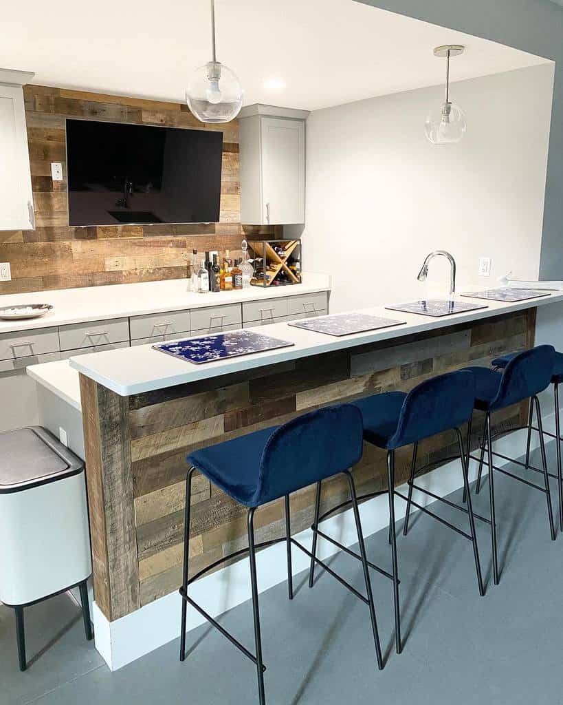 Modern rustic wet bar with reclaimed wood paneling, white countertop, and navy upholstered stools.