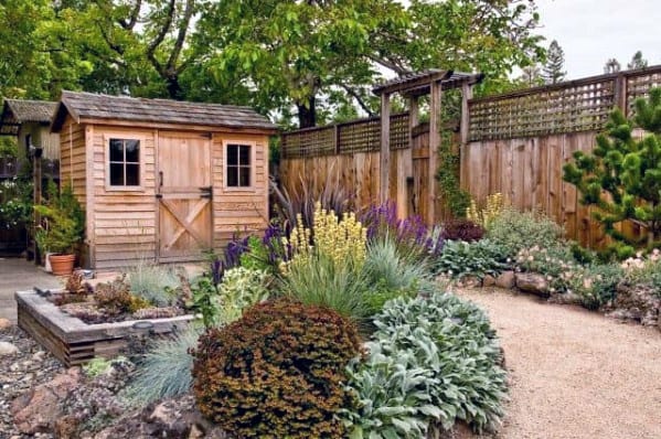 Rustic wooden shed with farmhouse-style doors in a garden with lush landscaping and flowers.