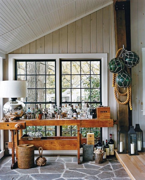 Rustic mini bar with wood table, bottles, nautical decor, and stone flooring by large windows.