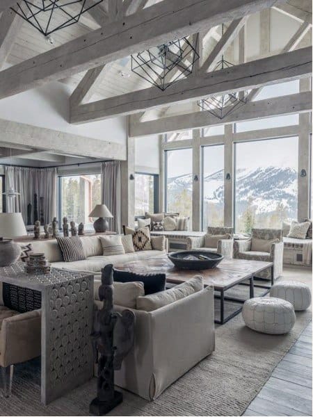 A rustic-themed living room with high ceilings, exposed wooden beams, large windows showcasing snowy mountain views, and elegant furniture, creating a serene and stylish space