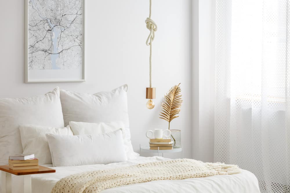 Minimalist white bedroom with white bedding, a hanging light bulb, and a decorative gold leaf accent on the side table