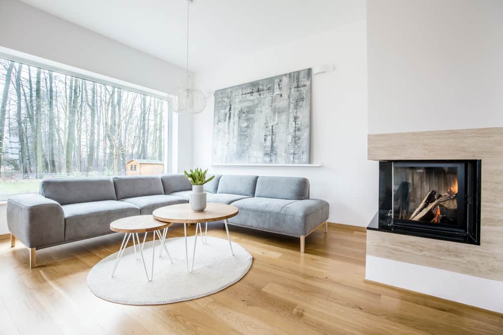 Scandinavian living room with a gray sectional sofa, wooden accents, a cozy fireplace, and a large window showcasing natural surroundings