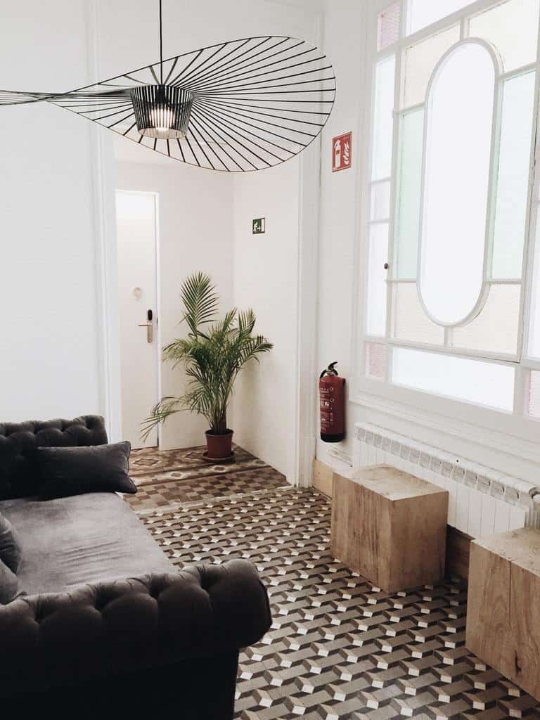 Minimalist living room with geometric tiled flooring, a tufted gray sofa, wooden stools, a potted plant, and a unique ceiling light fixture