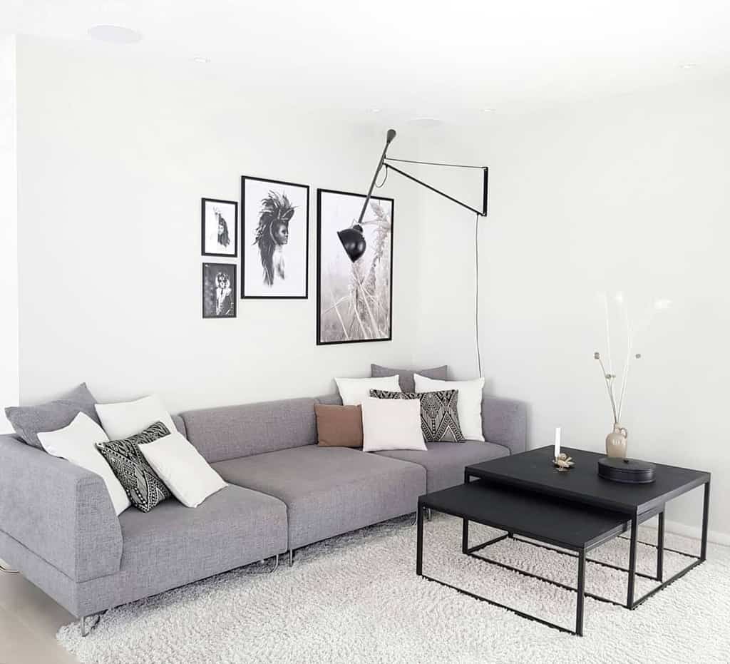 Minimalist living room with a gray sectional sofa, black nesting tables, monochrome wall art, and a sleek modern wall lamp on a white backdrop